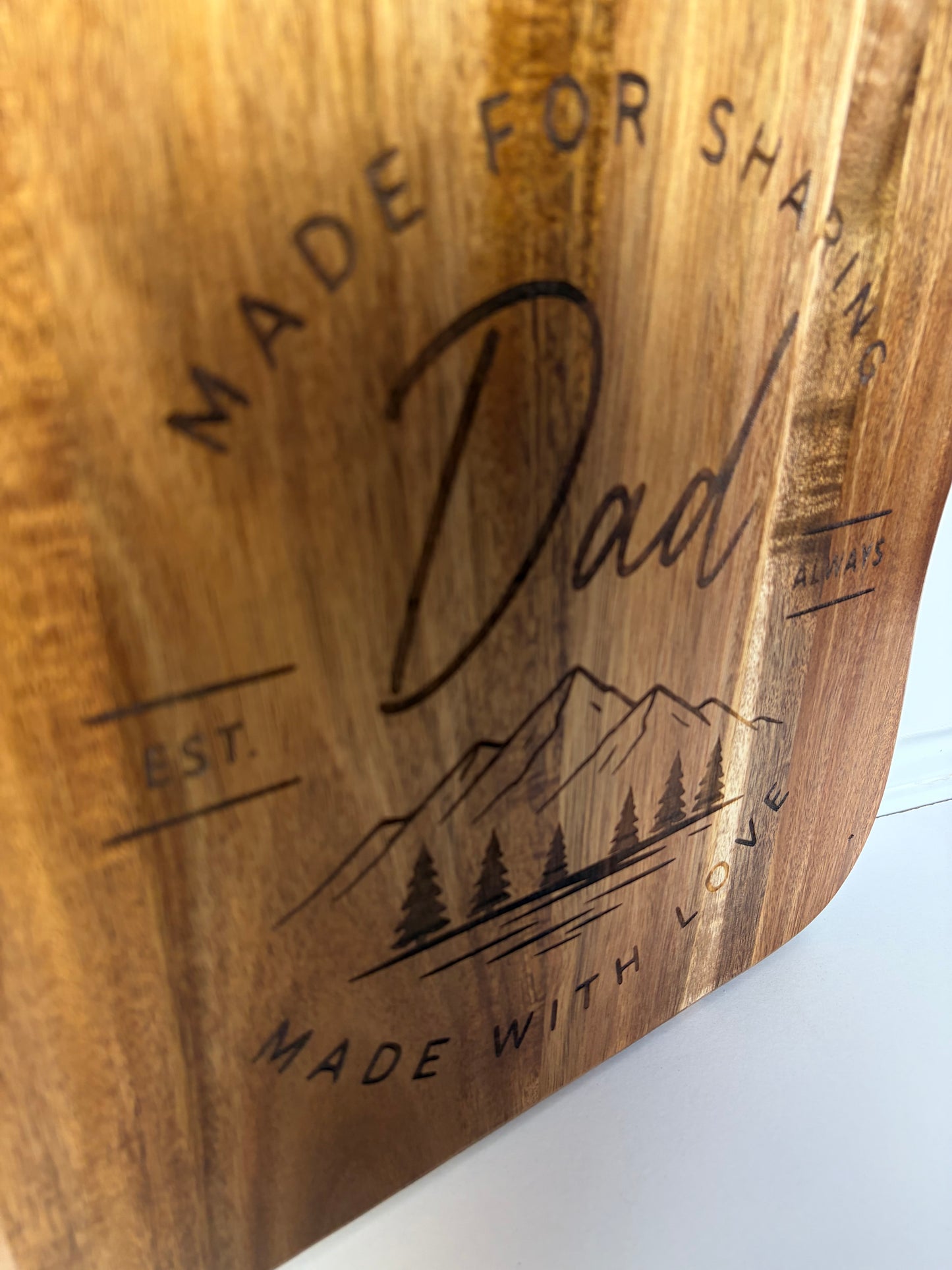 A close-up of a wooden SERVING BOARD with 'Dad', 'Made for sharing', 'Always', 'Made with love', and a mountain-and-trees illustration, shown on a white surface.