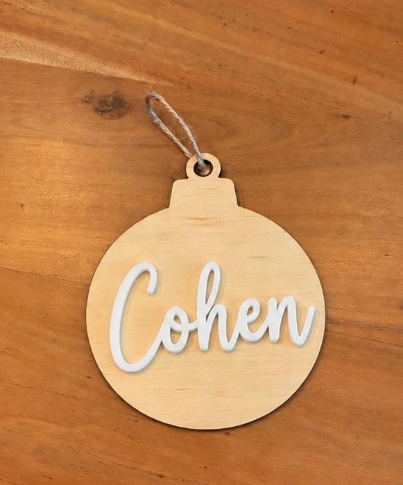A round natural wood ornament hanging on a wooden surface, with the name 'Cohen' cut out in white script across the front.