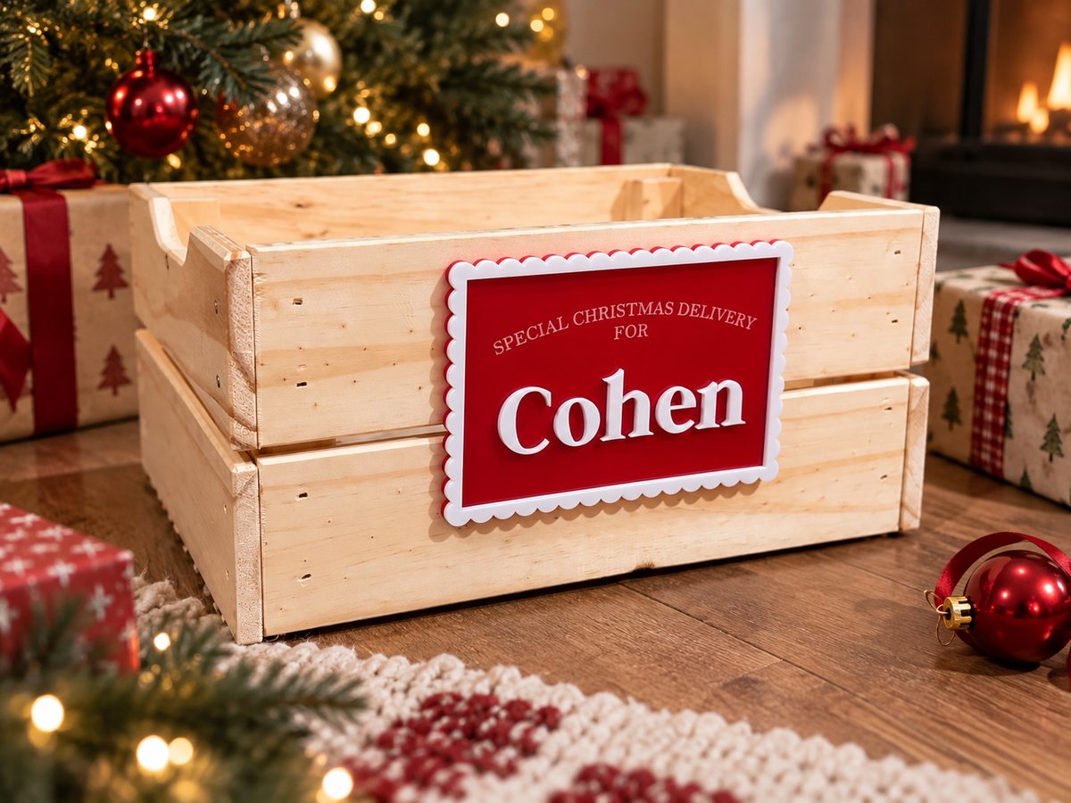 A wooden crate with a red personalized Christmas sign reading 'Cohen' and 'Special Christmas Delivery For' in white text, styled on a wooden floor with Christmas decorations, wrapped gifts, and a fireplace in the background.