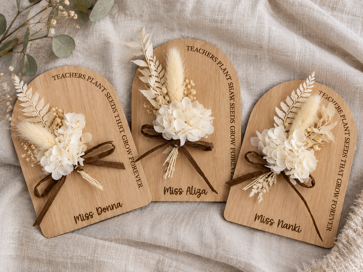 Three personalized wooden plaque favors for teachers, decorated with cream dried flowers, pampas grass, and brown ribbons, arranged on a beige linen fabric background.