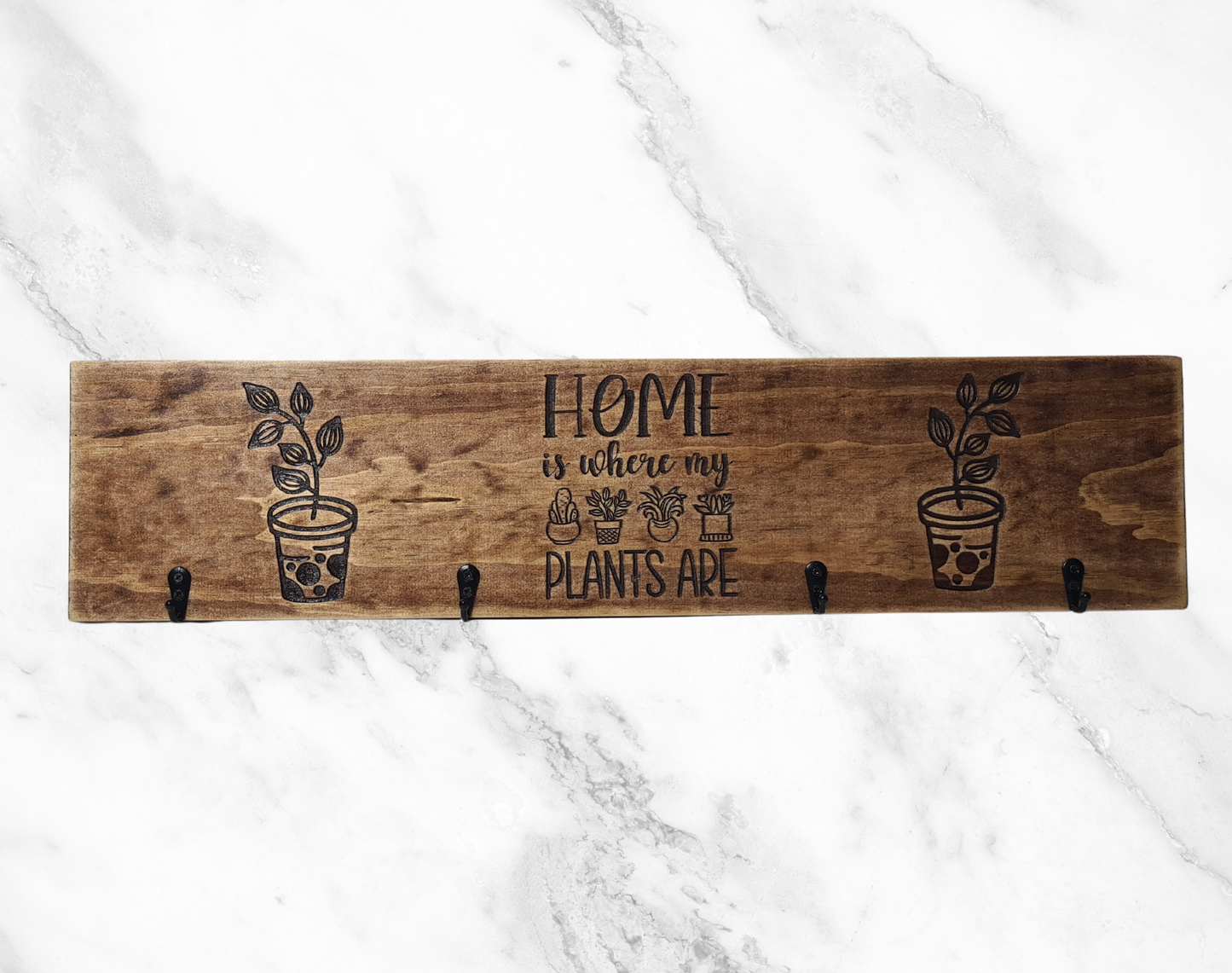 A wooden keyholder with four hooks, each decorated with a plant design and the phrase 'Home Is Where My Plants Are' engraved above them. The keyholder is displayed against a marble background.