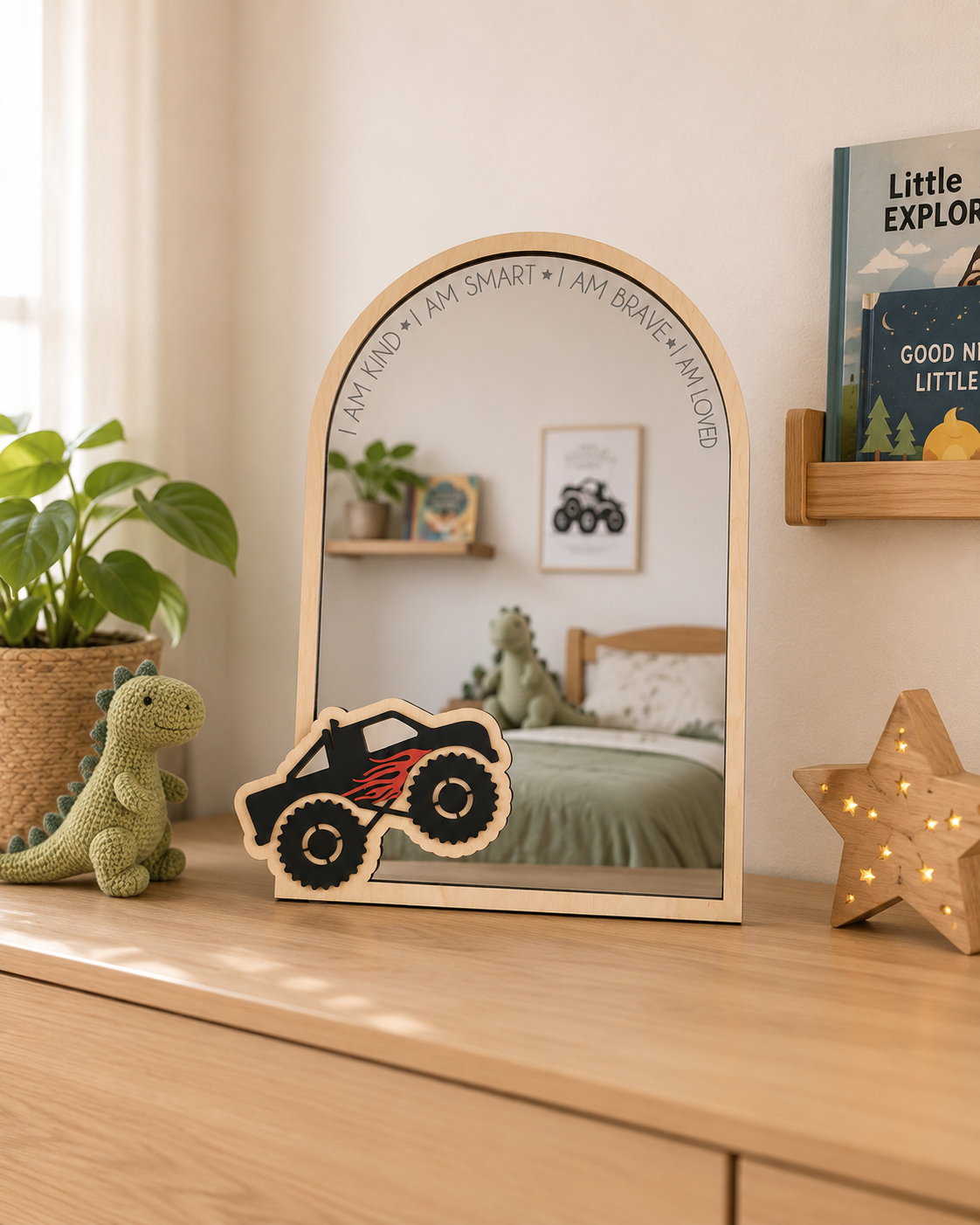 A wooden arch-shaped mirror with a small monster truck cutout in front of it, styled on a light wood dresser in a child's bedroom with a plant, book, and star lamp visible.