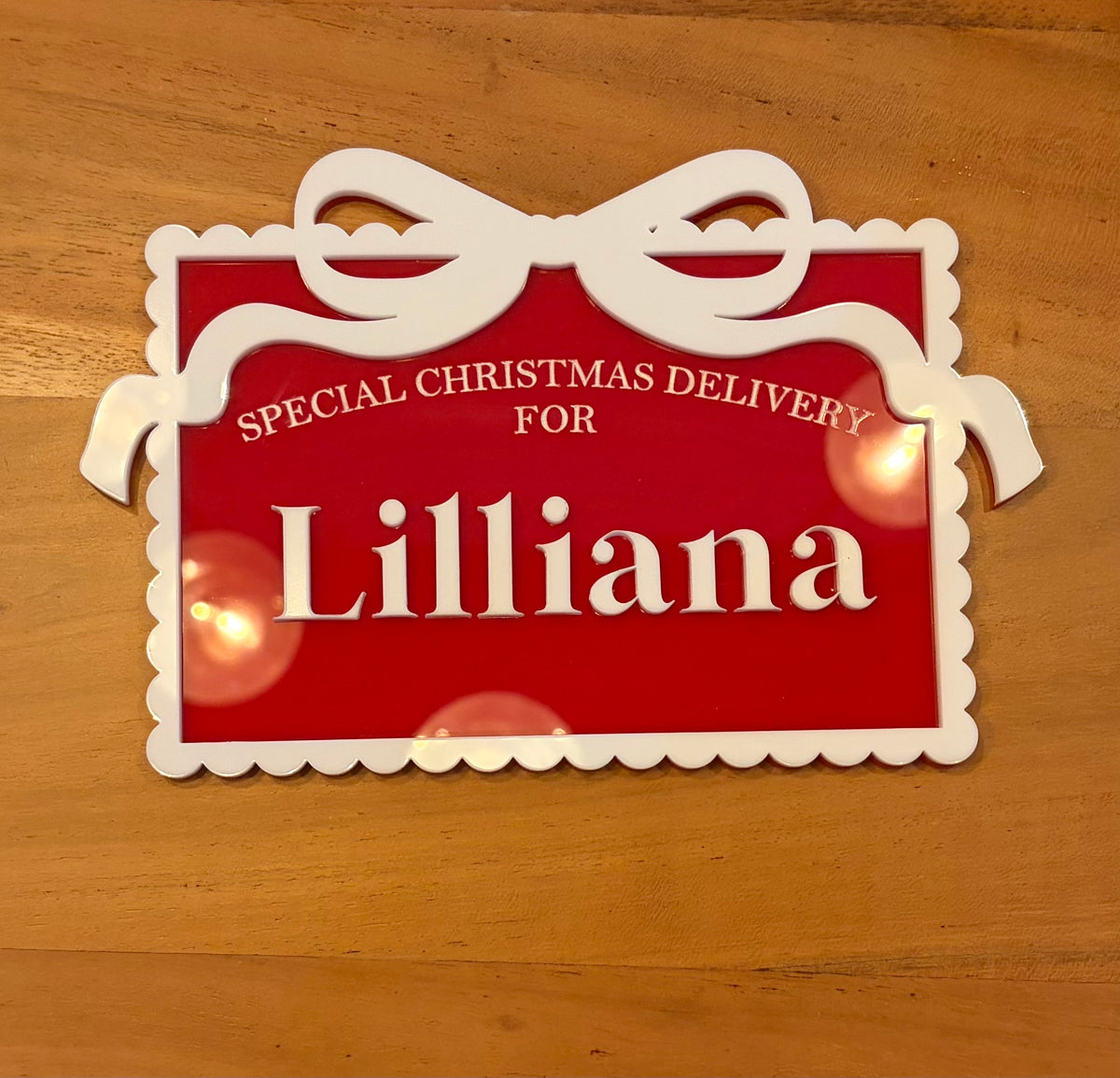 A red and white personalized Christmas sign with the text 'SPECIAL CHRISTMAS DELIVERY FOR Lilliana' on a wooden tabletop.
