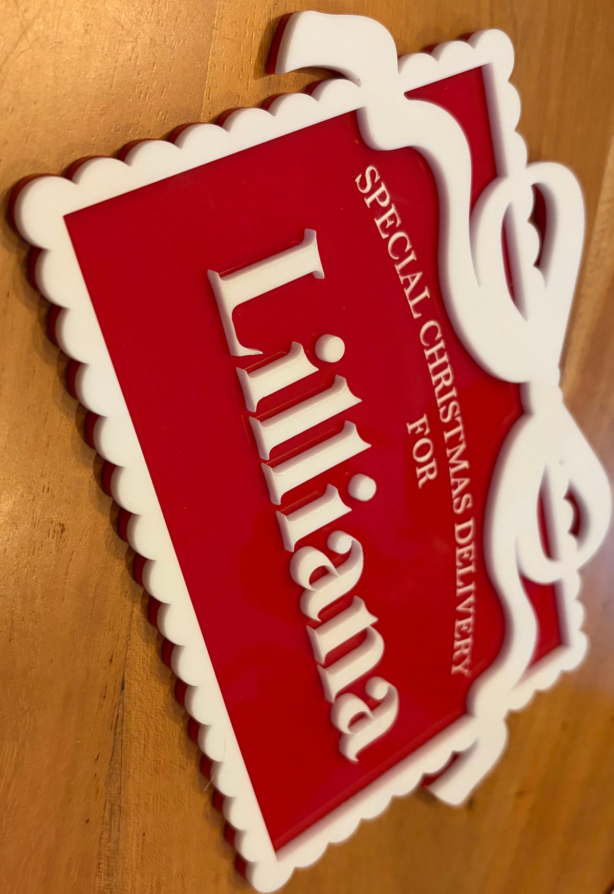 A red and white decorative sign with the name 'Liliana' in large white letters and the text 'SPECIAL CHRISTMAS DELIVERY FOR' printed on it, photographed on a wooden surface.