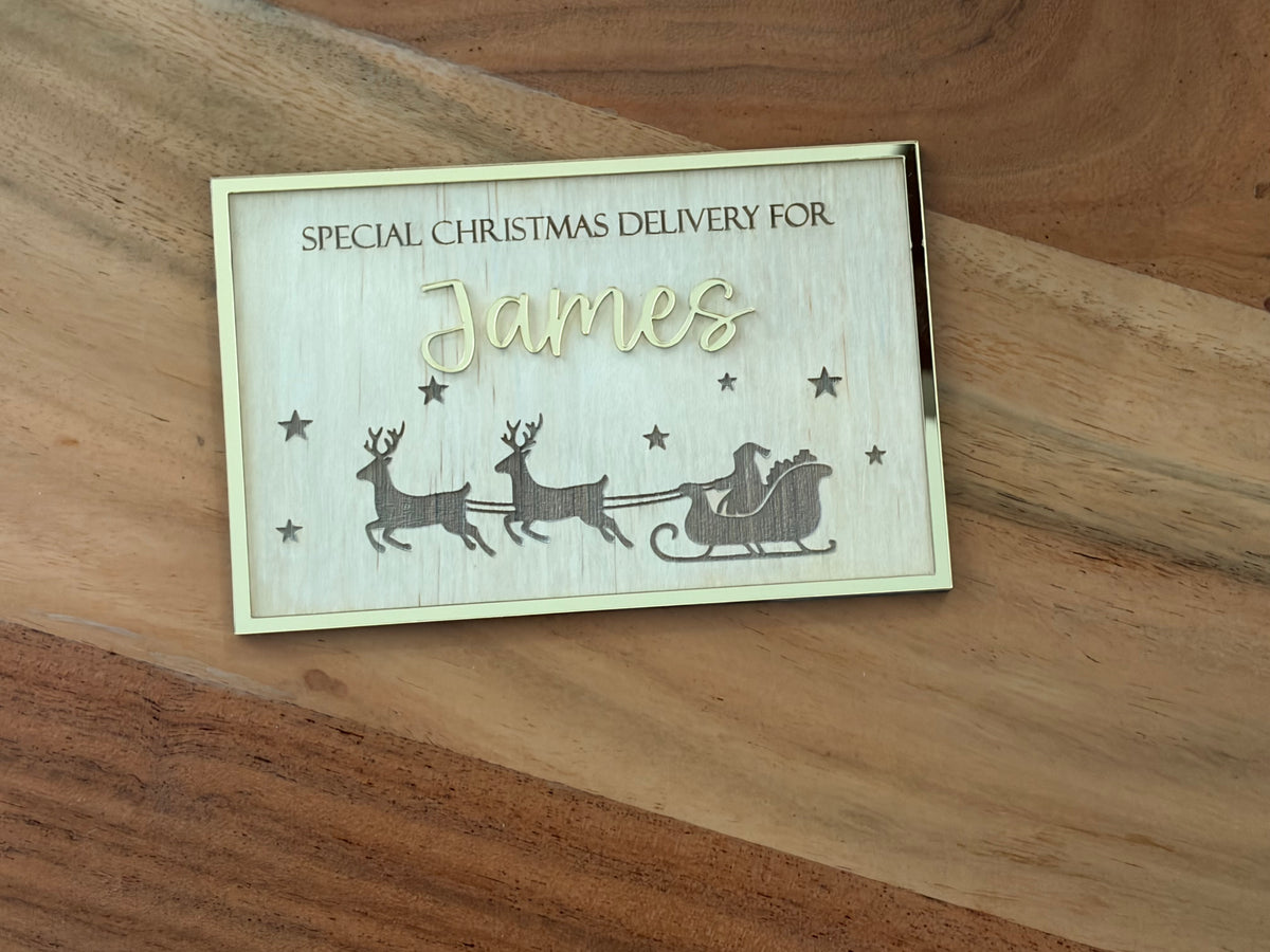 A rectangular personalized wooden Christmas gift box lid or tag with laser-engraved text reading 'SPECIAL CHRISTMAS DELIVERY FOR James' and a holiday scene with reindeer, Santa, and stars, placed on a wooden surface.