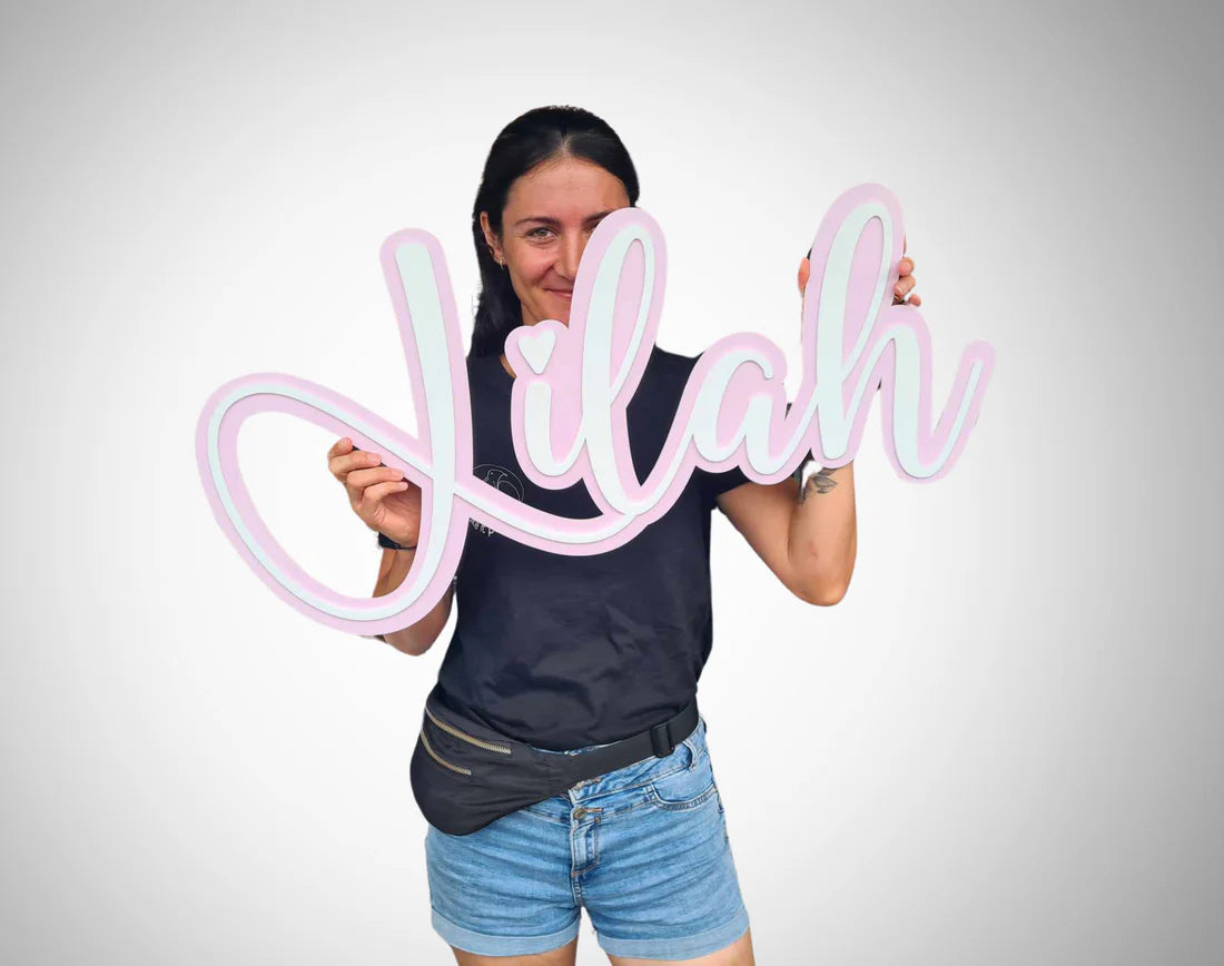 A woman holding a large pink and white script sign that says 'Lilah' in front of her face, wearing a black t-shirt, denim shorts, against a light gray gradient background.