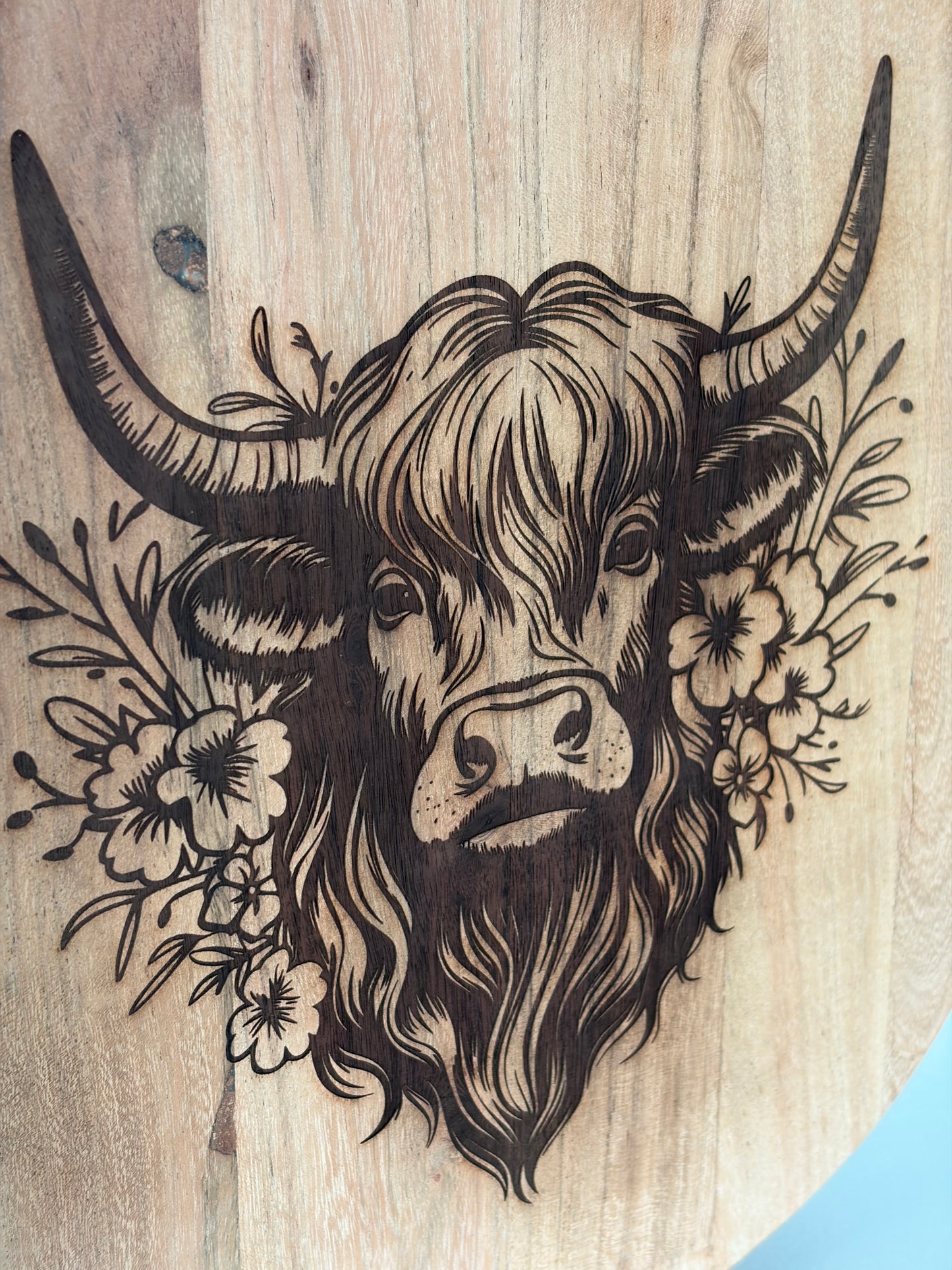 A close-up of a wooden surface featuring a detailed black-burned illustration of a highland cow with long horns and a floral crown, set against a light natural wood grain background.