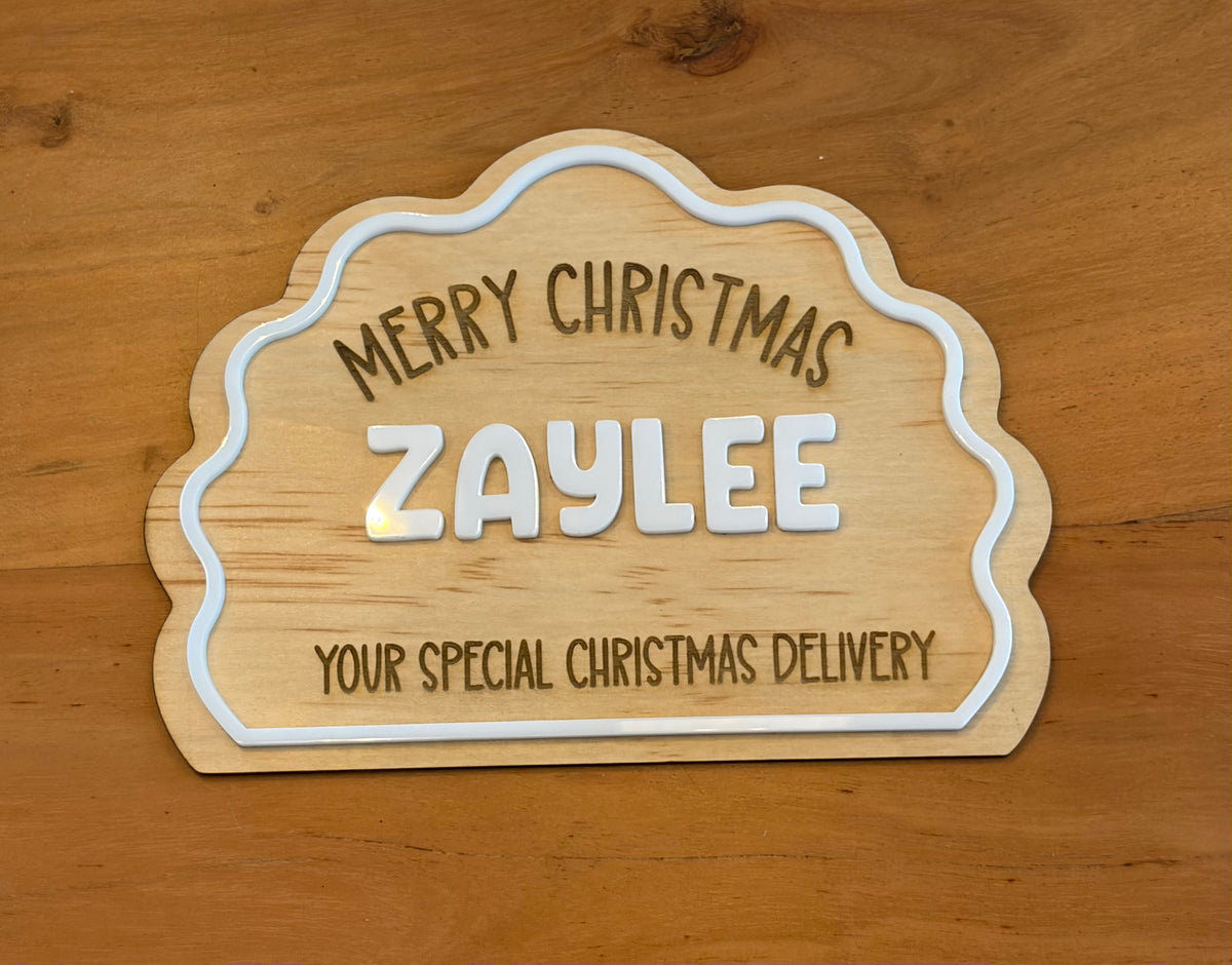A personalized wooden Christmas sign with a scalloped edge, featuring the text 'MERRY CHRISTMAS ZAYLEE YOUR SPECIAL CHRISTMAS DELIVERY' in dark brown and white lettering, displayed on a wooden tabletop.