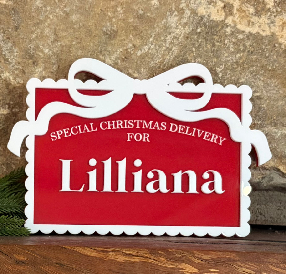 A red and white personalized Christmas gift box sign with the text 'SPECIAL CHRISTMAS DELIVERY FOR Lilliana' in white lettering, displayed on a wooden surface against a textured stone background.