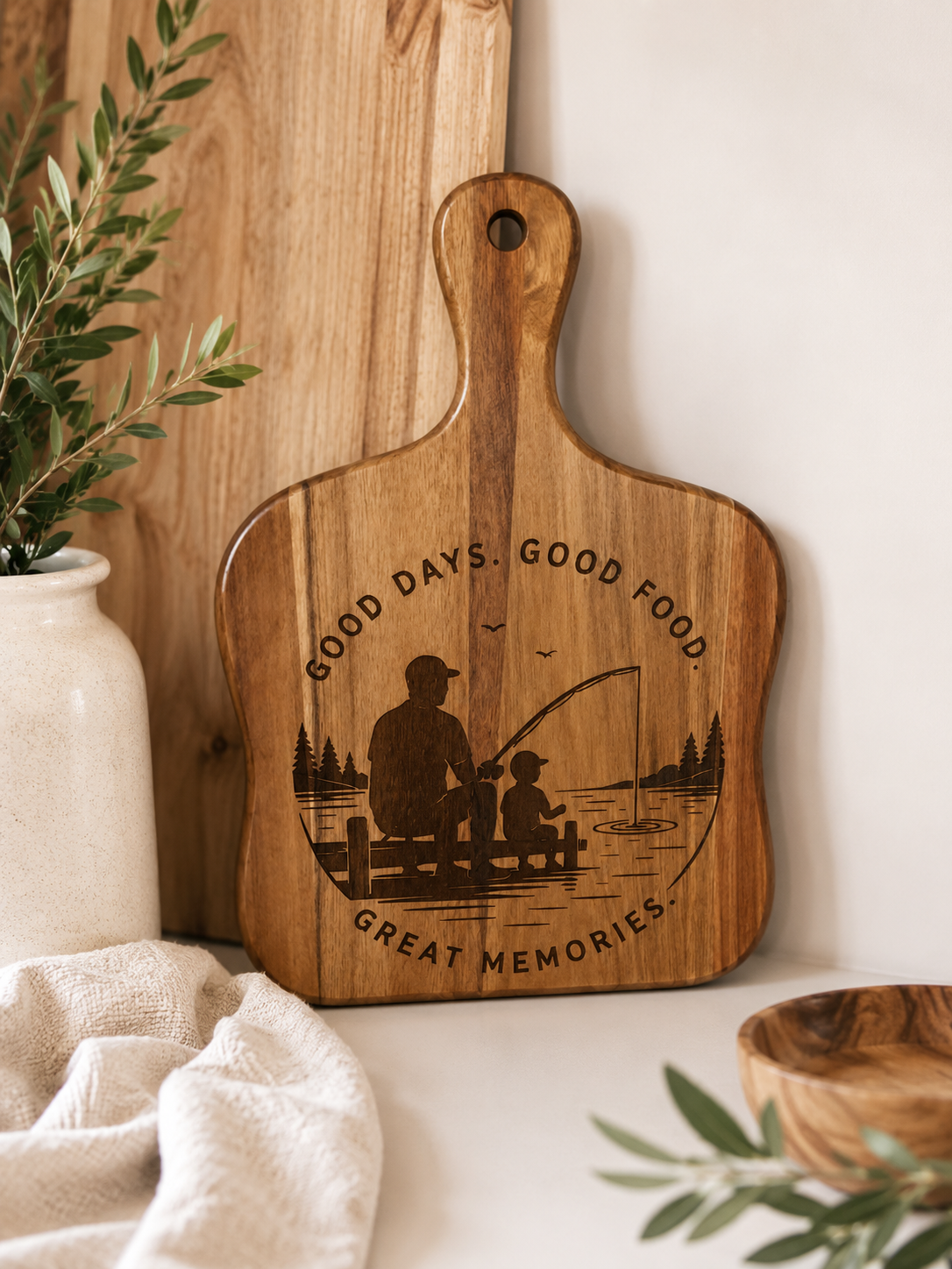 A wooden cutting board with a handle, featuring an engraved design of a father and child fishing on a dock with the text 'GOOD DAYS. GOOD FOOD. GOOD MEMORIES'