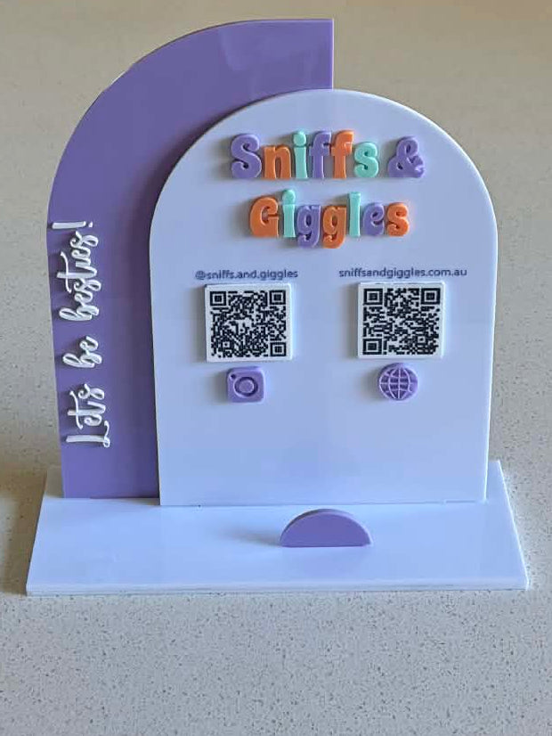 A small freestanding display stand shaped like a house, placed on a light countertop in a real indoor room with furniture and curtains visible in the background. The stand is white and lavender with colorful text reading 'Biblical Living' and QR codes on the front.