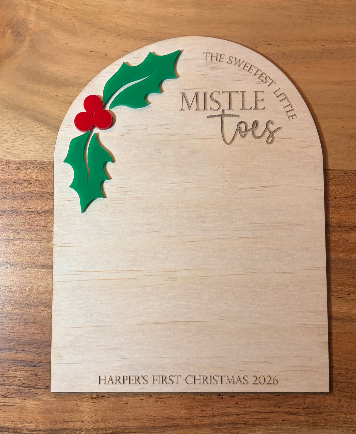 A personalized wooden Christmas plaque with an arched top, engraved with 'THE SWEETEST LITTLE MISTLE TOES' and 'HARPER'S FIRST CHRISTMAS 2026', decorated with a green holly leaf and red berries on a wooden surface.