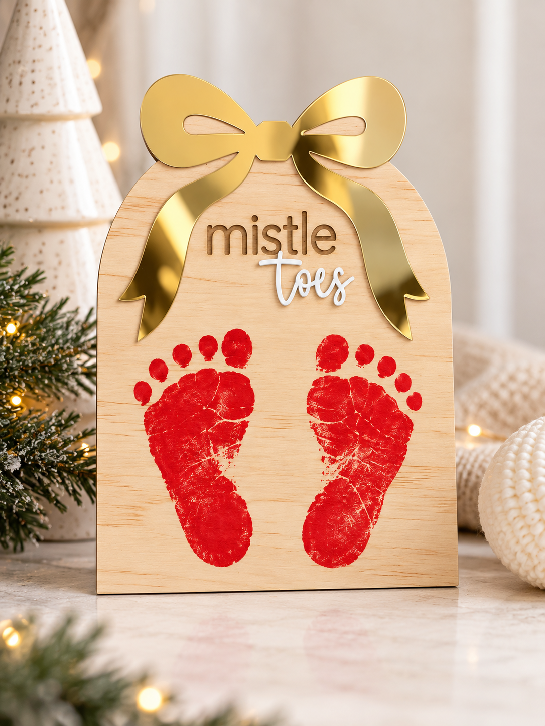 A personalized wooden gift tag plaque with a gold bow, the words 'mistle toes,' and two red baby footprints, styled on a tabletop with holiday greenery and warm lights in the background.