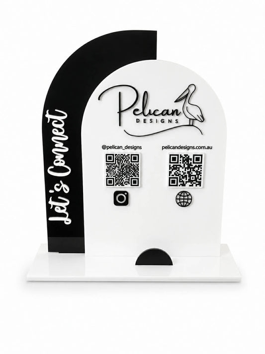 A custom acrylic tabletop sign for Pelican Designs, featuring a white arched main panel with the brand name, social handle, website, and QR codes, alongside a black side panel with the text 'Let's Connect'.