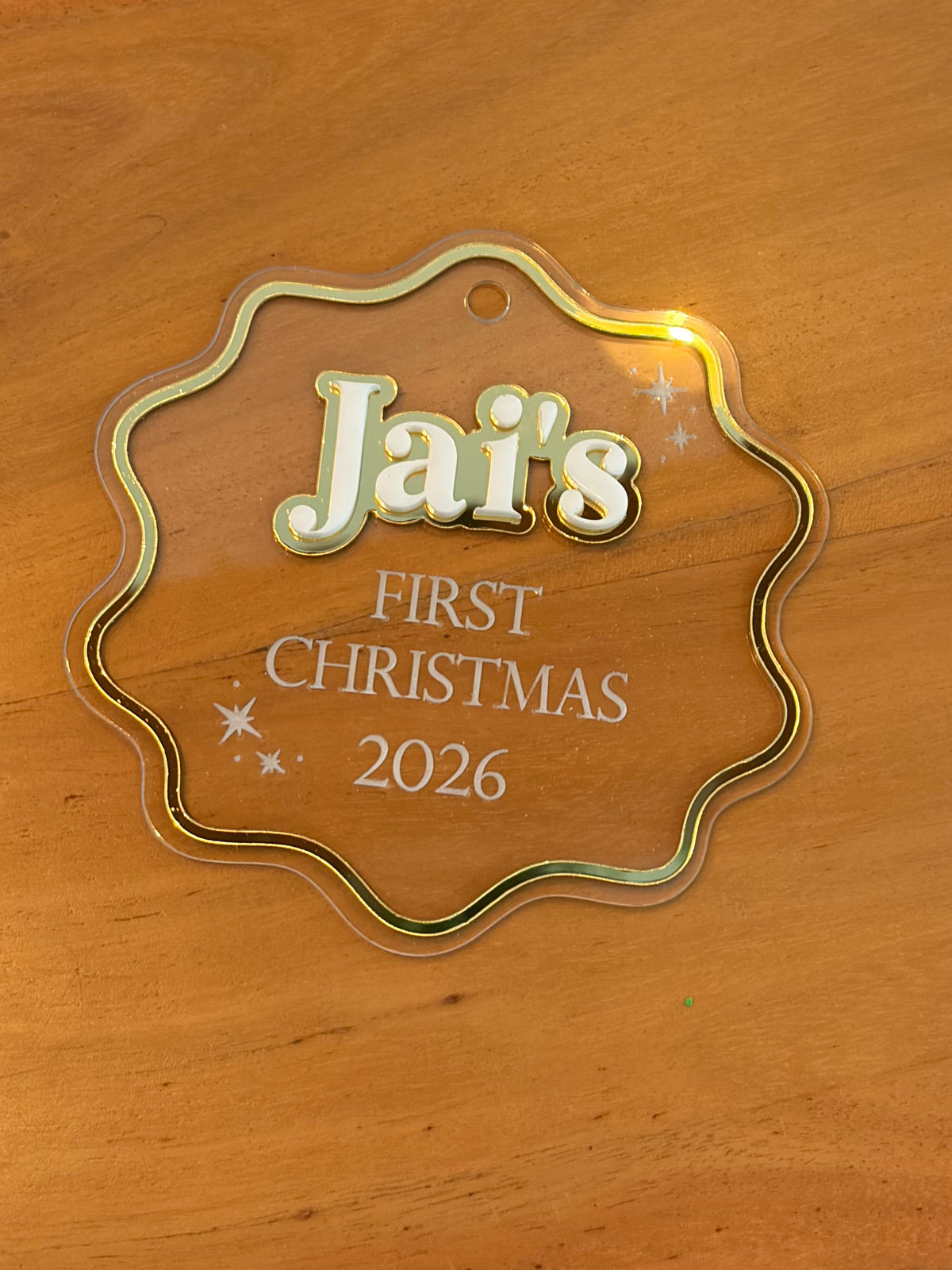A scalloped clear acrylic Christmas ornament with a gold-toned border and personalized text reading 'Jai's FIRST CHRISTMAS 2026' on a wooden tabletop.