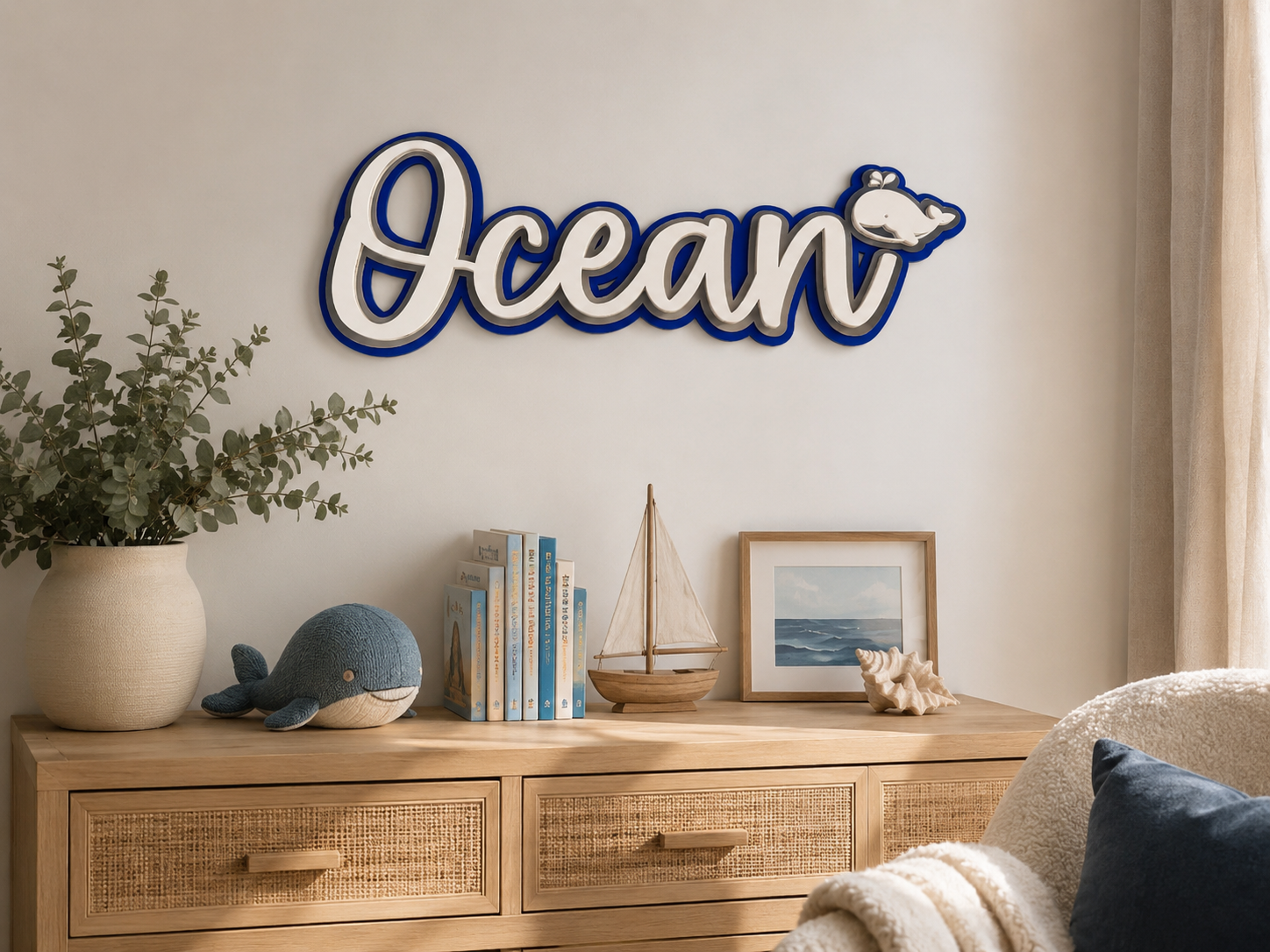 A decorative wall sign spelling 'Ocean' in white script with a dark blue border and a small whale detail, mounted above a wooden console table in a styled room with books, a sailboat model, a framed print, and a plant.