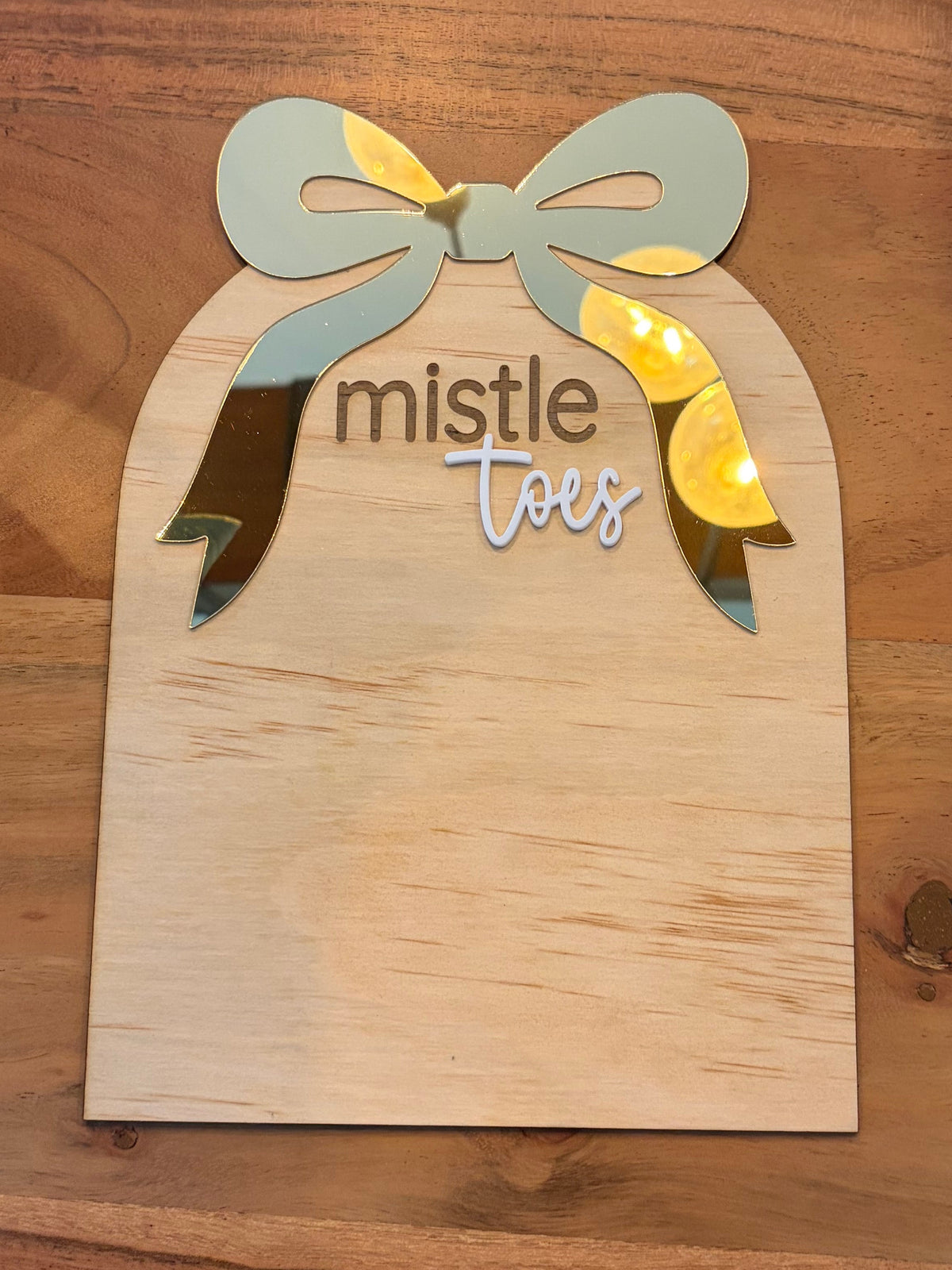 A wooden bell-shaped sign or plaque on a wooden table, decorated with a bow and the words 'mistle toes' in dark brown and white script.