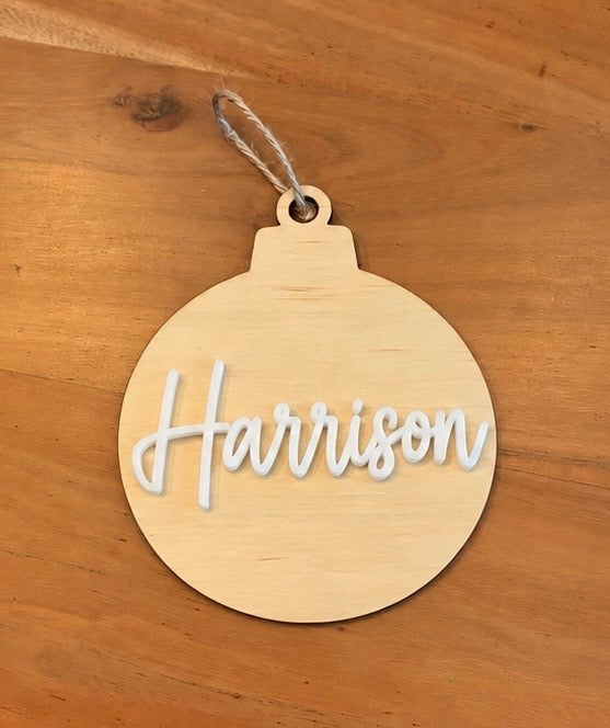A round wooden ornament with the name 'Harrison' cut out in white script, hanging from a twine string on a wooden surface.