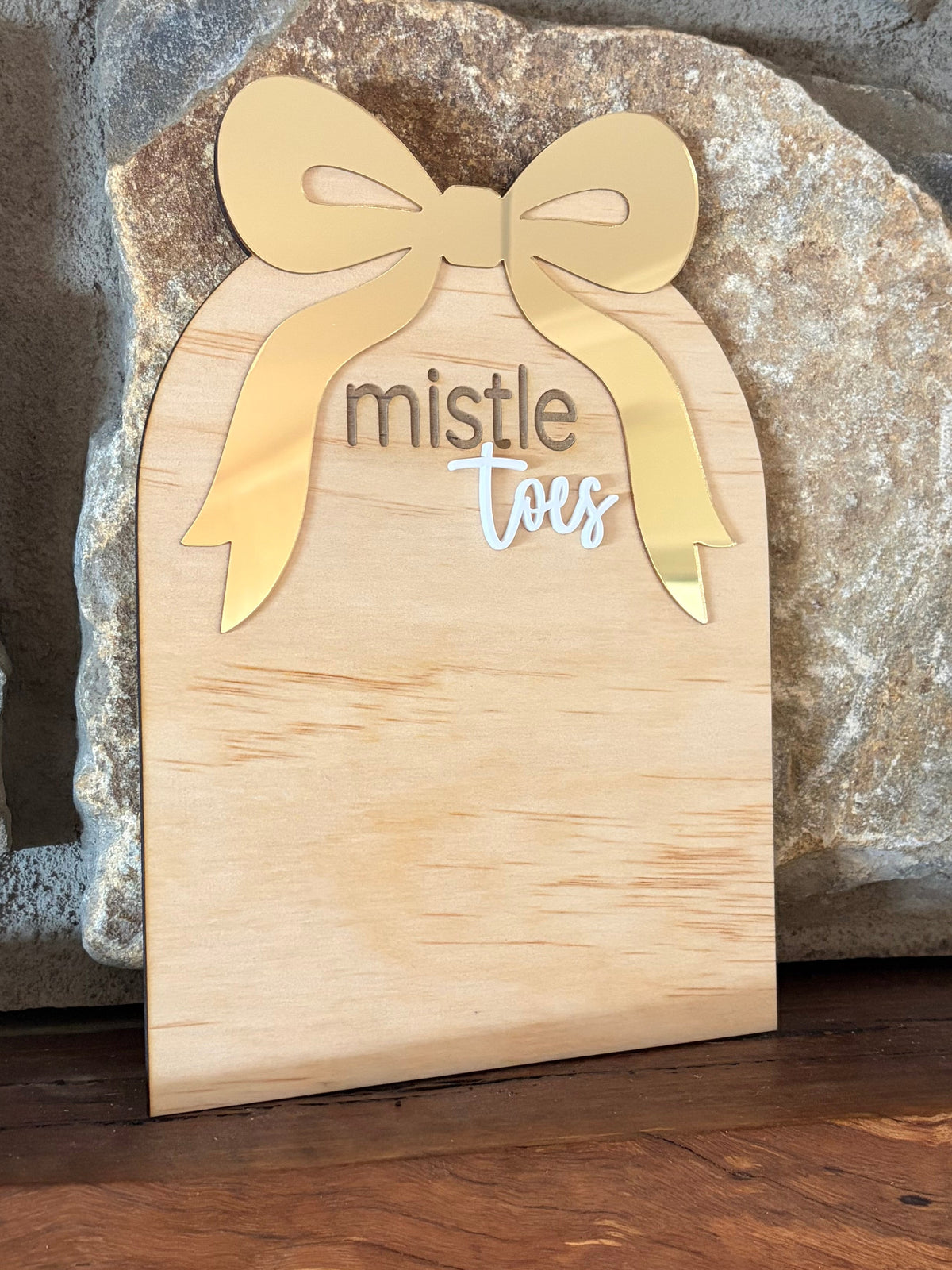 A small wooden gift box sign with a bow design and the words 'mistle toys' engraved, leaning against a stone wall on a wooden surface.