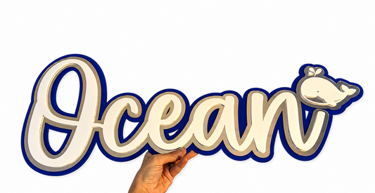 A hand with a floral tattoo holds a decorative 3D sign spelling 'Ocean' in a cursive font, featuring a small whale detail on the letter 'n'. The sign has a white face with a dark blue border and a silver mirror shadow layer, set against a plain white background.