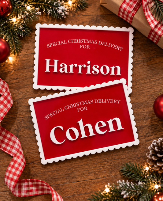 Two red Christmas-themed cards with white scalloped borders reading 'Special Christmas Delivery for Harrison' and 'Special Christmas Delivery for Cohen,' arranged on a wooden surface with holiday decorations including pine branches, lights, ornaments, ribbon, and a wrapped gift.