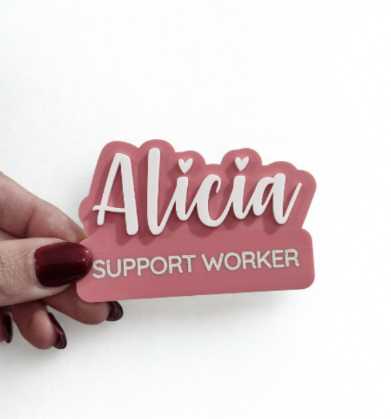 A hand with dark red nail polish holds a custom pink and white name sign that reads 'Alicia SUPPORT WORKER' against a plain white background.