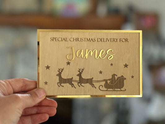 A hand holds a rectangular wooden Christmas gift box with a gold laser-engraved design featuring Santa, reindeer, stars, and the text 'SPECIAL CHRISTMAS DELIVERY FOR James'.