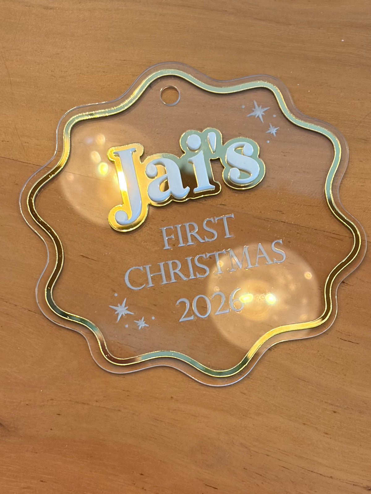 A scalloped clear acrylic hanging ornament with a holographic border and personalized text reading 'Jai's FIRST CHRISTMAS 2026' on a wooden surface.