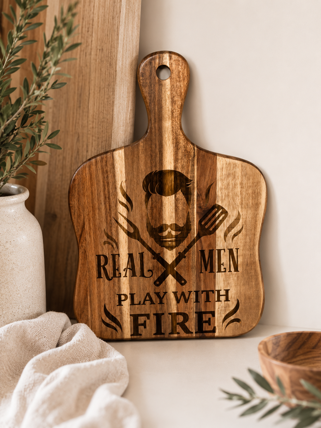 A wooden cutting board with a handle, featuring a laser-engraved graphic of a bearded man with crossed spatulas and the text 'REAL MEN PLAY WITH FIRE'. The cutting board is styled in a warm kitchen setting next to a ceramic vase with greenery and a beige cloth.