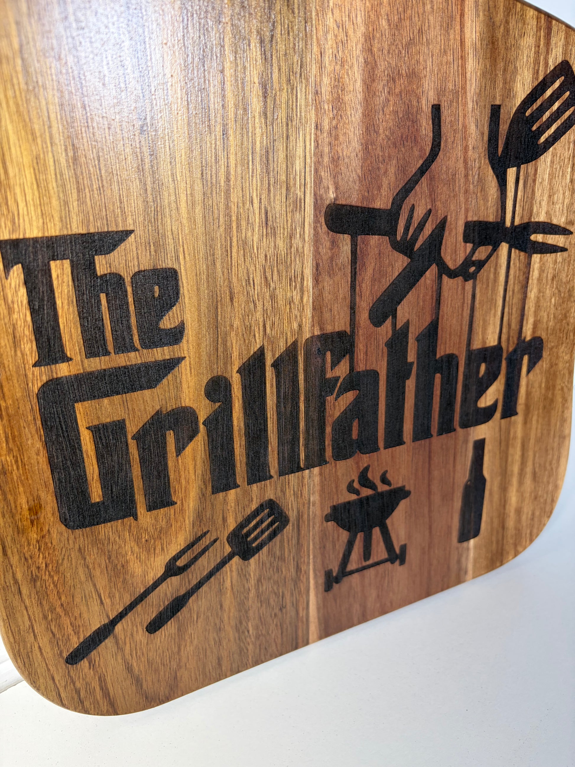 A close-up of a wooden cutting board with rounded corners, featuring a black engraved design that reads 'The Grillfather' with barbecue tools and a grill.