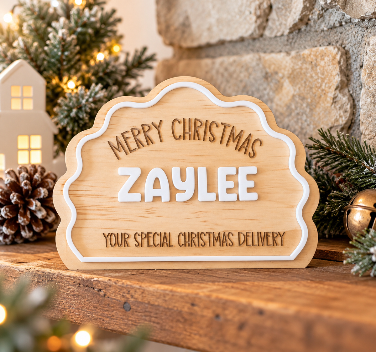 Personalized wooden Christmas sign with scalloped edges, engraved text reading 'MERRY CHRISTMAS ZAYLEE YOUR SPECIAL CHRISTMAS DELIVERY,' displayed on a rustic wooden surface with holiday greenery, a stone wall, and warm lights in the background.