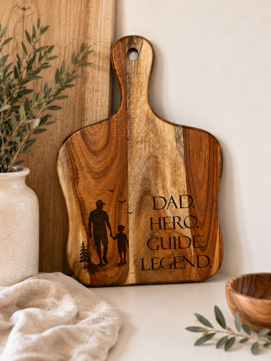 A wooden cutting board with a handle, featuring engraved black silhouettes of a father and child and the words 'DAD HERO GUIDE LEGEND'. The board is styled on a tabletop with a neutral background, a ceramic vase with greenery, and a linen cloth.