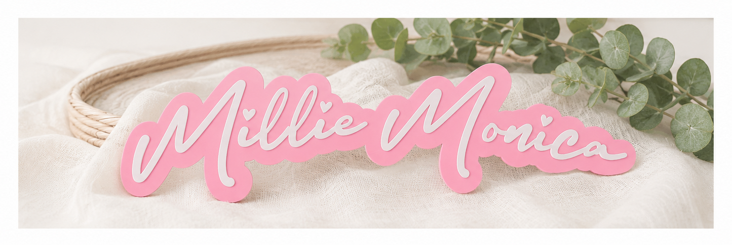 Pink acrylic script sign reading 'Millie Monica' shown in two views: a standalone top view and a styled bottom view draped over a rope and green eucalyptus leaves on a light fabric background.