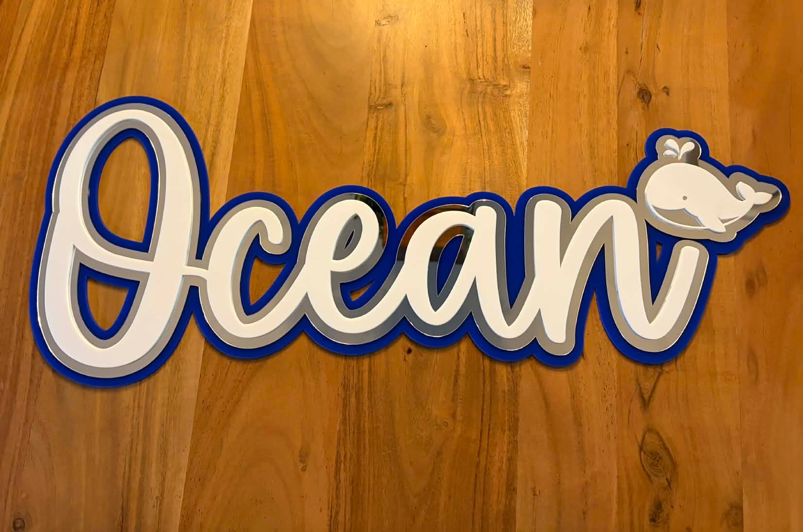 A decorative wall sign spelling 'Ocean' in white cursive letters with a blue outline, plus a small white whale graphic at the upper right, shown against a wood plank background.