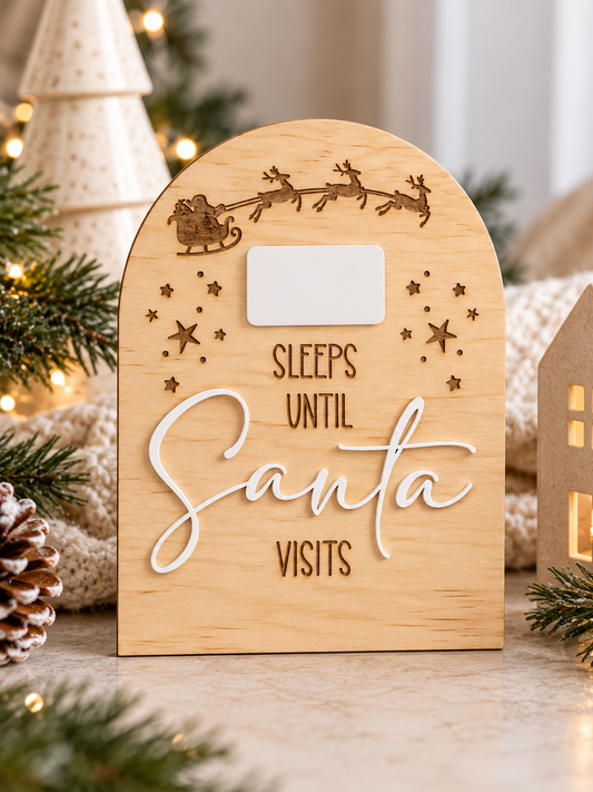 A wooden arched sign with laser-engraved Santa and sleigh design, featuring the phrase 'SLEEPS UNTIL Santa VISITS' in white script and block letters, with a blank white rectangular area near the top. The sign is styled on a tabletop with holiday greenery, a pinecone, and warm string lights in the background.