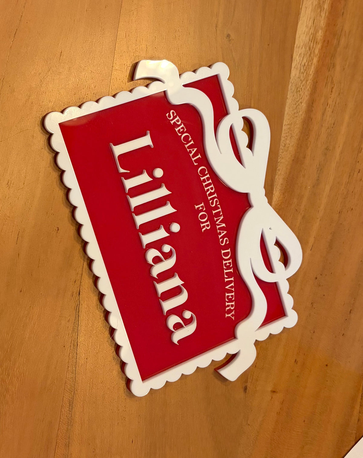A red and white scalloped Christmas gift tag sign with the name 'Lilliana' in white script, placed on a wooden surface. The sign includes the text 'SPECIAL CHRISTMAS DELIVERY'.