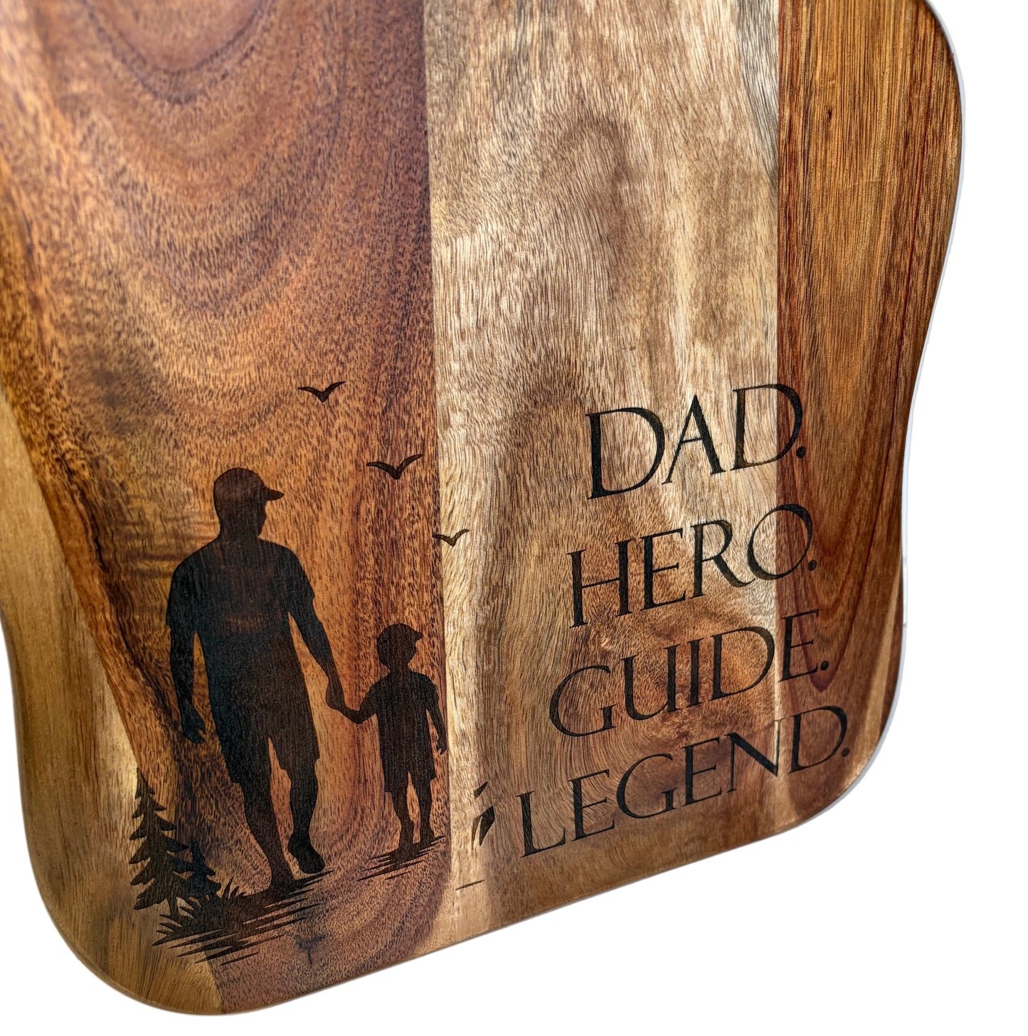 A close-up of a wooden serving board with rounded corners, featuring natural wood grain, engraved silhouettes of a man and child walking with trees and birds, and the words 'DAD HERO GUIDE LEGEND' on a white background.