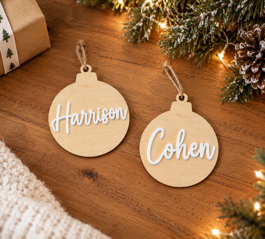 Two personalized wooden Christmas ornaments with the names 'Harrison' and 'Cohen' in white script, displayed on a wooden tabletop with holiday decorations and warm string lights.