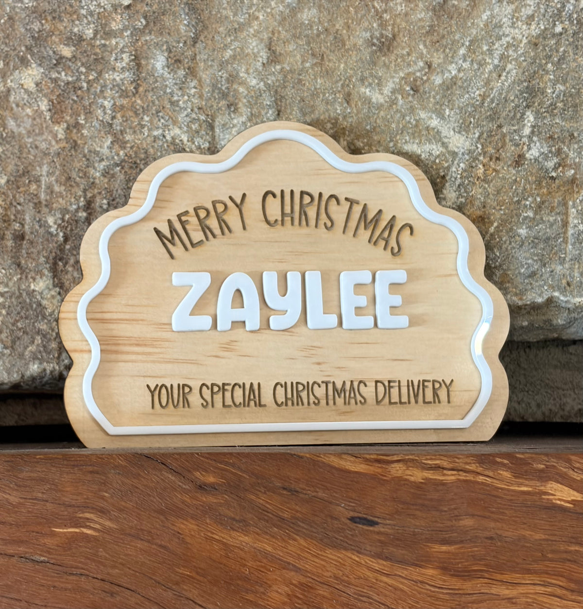 A scalloped wooden Christmas sign with white lettering that reads 'MERRY CHRISTMAS ZAYLEE YOUR SPECIAL CHRISTMAS DELIVERY', displayed against a stone wall and wood surface.