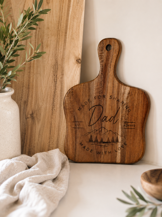 A wooden cutting board with a handle, engraved with 'Dad' and mountain graphics, styled on a tabletop with a neutral cloth, a wooden panel, and greenery.