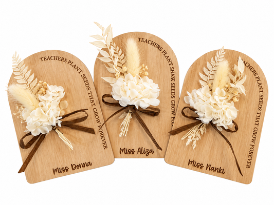Three personalized wooden plaque ornaments shaped like arched signs, each decorated with cream dried flowers, beige grasses, and a brown ribbon, with engraved names and the phrase 'Teachers plant seeds that grow forever.'.