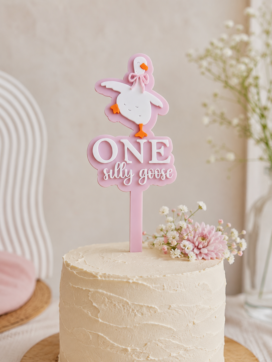Pink personalised One Silly Goose acrylic cake topper displayed on a neutral buttercream birthday cake with soft floral styling for a first birthday celebration.