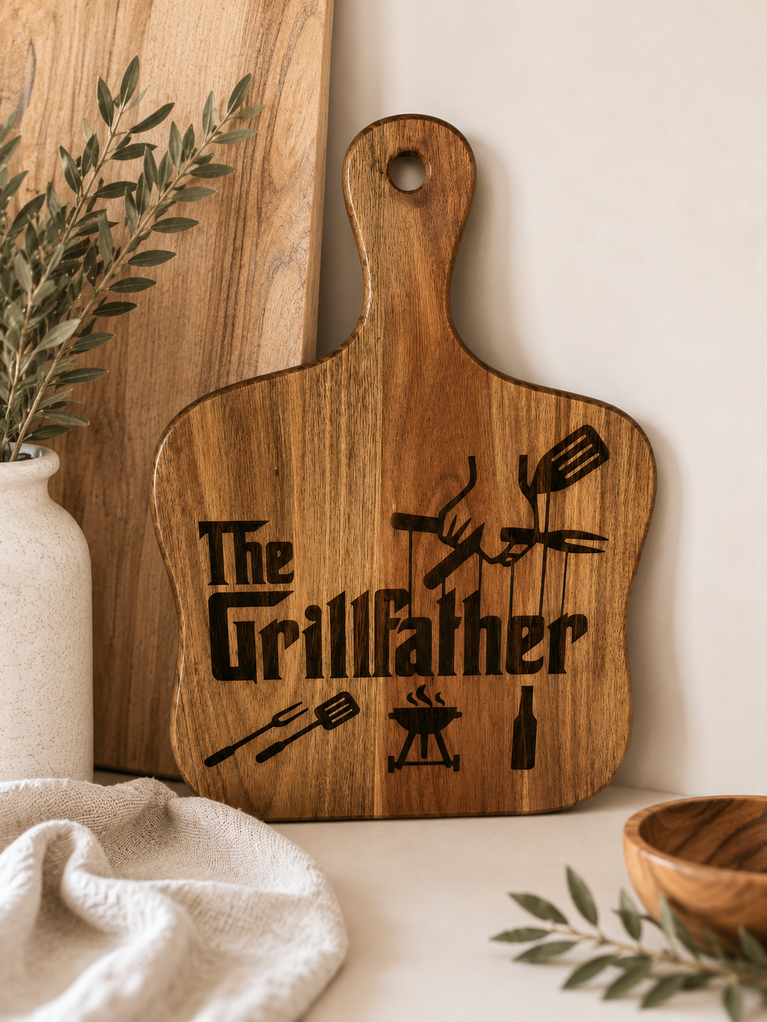 A wooden cutting board with a handle, featuring the engraved text 'The Grillfather' and grill-themed utensil illustrations, styled in a kitchen setting.