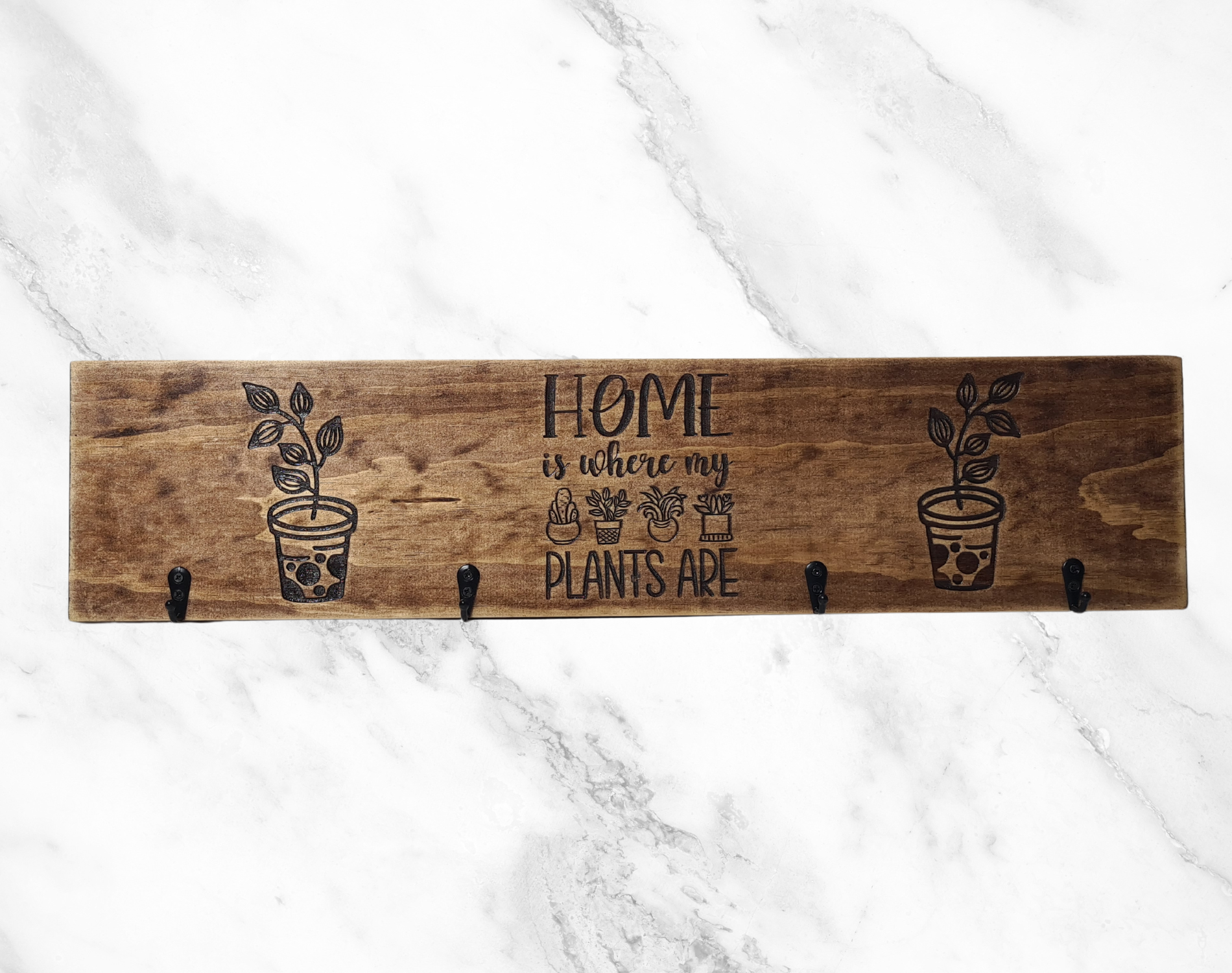 A wooden keyholder with four hooks, each decorated with a plant design and the phrase 'Home Is Where My Plants Are' engraved above them. The keyholder is displayed against a marble background.