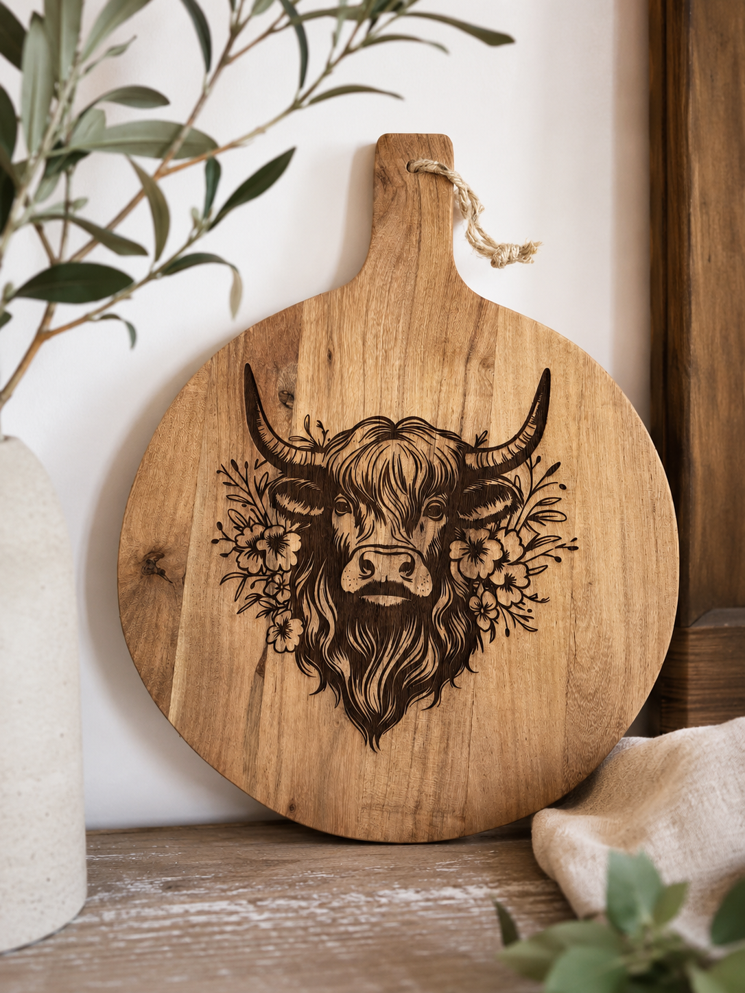 A round wooden cutting board with a handle, featuring a dark engraved illustration of a highland cow surrounded by flowers. The board is styled on a rustic wooden table next to a white vase with green leaves and a beige cloth.