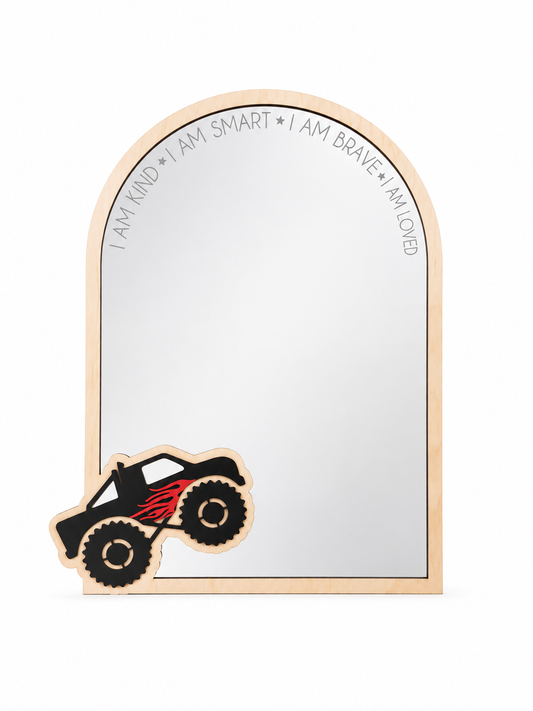 A wooden arch-shaped mirror with a light natural wood frame, featuring the words 'I AM KIND • I AM SMART • I AM BRAVE • I AM LOVED' printed along the top curve. A matching wooden monster truck decoration with black wheels and red flame decals rests in the bottom-left corner on a clean white background.
