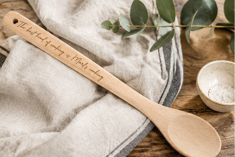 A wooden spoon with engraved text is shown on  a linen cloth with a small bowl and greenery nearby. The engraving reads, 'The best kind of cooking is Marl's cooking.'