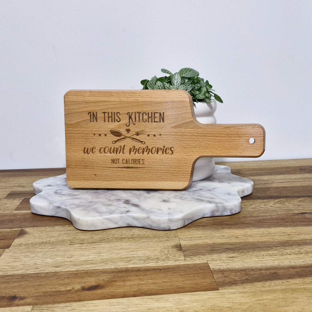 An engraved bamboo serving board with the phrase 'In this kitchen we count memories, not calories' printed on it, placed on a marble surface with a mint plant in the background.
