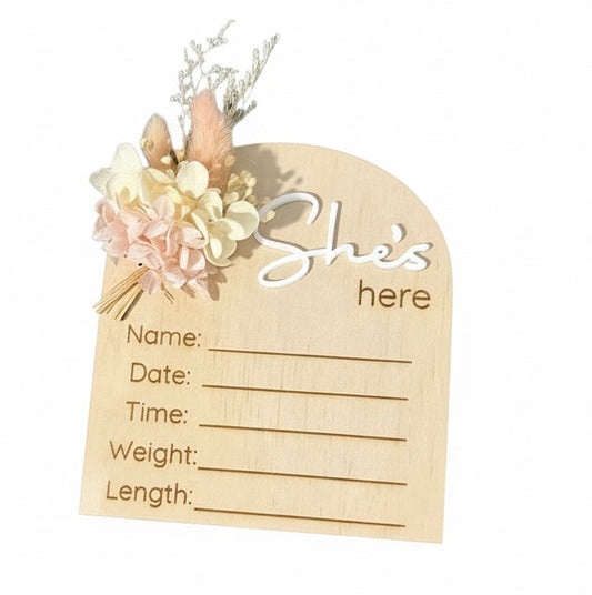 Baby announcement arch made from plywood with floral decoration with text she's here on a white background